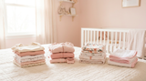 A neatly folded stacks of newborn baby clothes — onesies, footed sleepers, swaddle blankets, and burp cloths — laid out on a soft cream bed cover in a blush pink nursery.