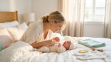 Mother smiling while dressing her newborn baby in a soft cotton onesie on a bed in warm natural light.